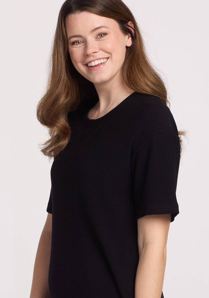 A young woman with long brown hair smiles at the camera, wearing the Woolx Vera Ribbed Tunic in black. She stands against a plain light background.