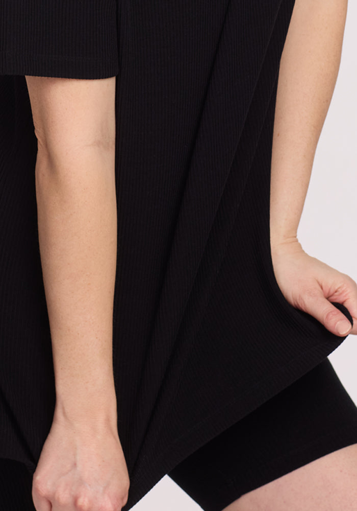 Close-up of a person wearing the Woolx Vera Ribbed Tunic in black with black shorts, gently pulling the shirt's side. The background is plain and light-colored.