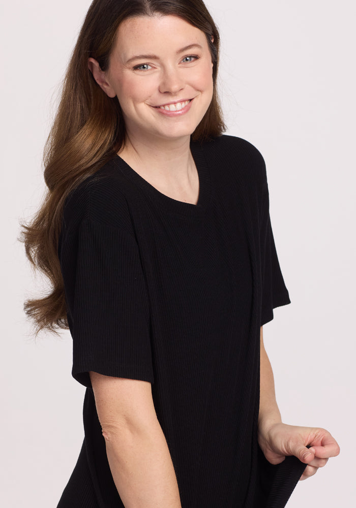 A woman with long brown hair smiles at the camera, wearing the Woolx Vera Ribbed Tunic in black. This relaxed fit top has short sleeves, and she holds its hem. The background is plain and light-colored.