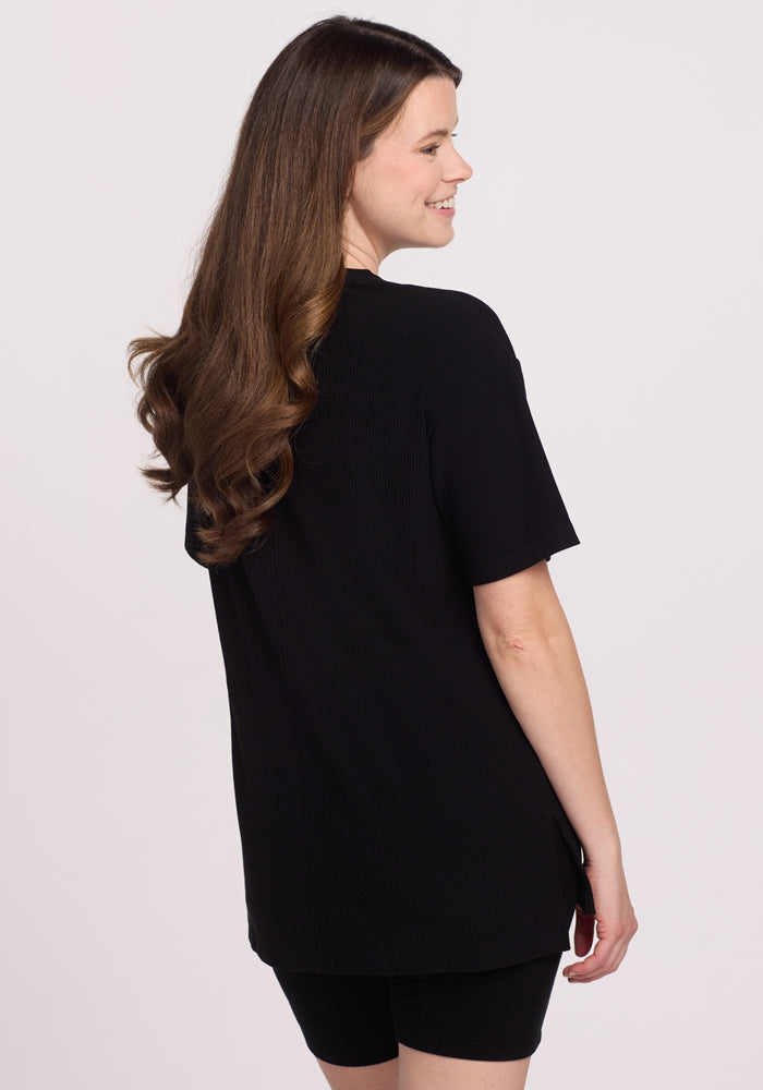 A woman with long, wavy brown hair smiles as she faces away from the camera, wearing the Woolx Vera Ribbed Tunic in black with matching black shorts against a plain light background.