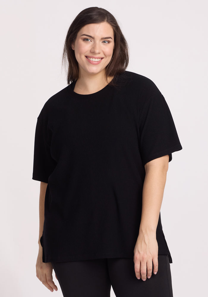 A woman with shoulder-length brown hair smiles at the camera, wearing the Woolx Vera Ribbed Tunic in black and matching pants, standing against a plain light background.