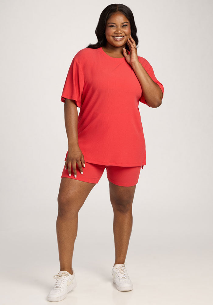 Smiling woman wears the Woolx Vera Ribbed Tunic in Hibiscus, paired with shorts and white sneakers against a plain background.