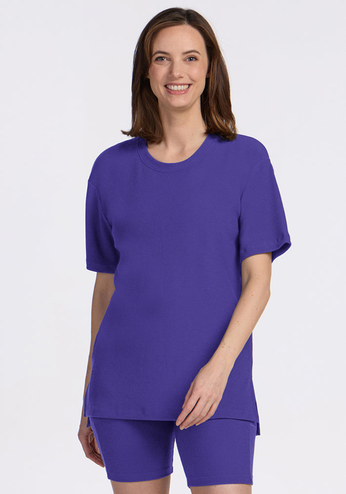 A smiling woman wears the Woolx Vera Ribbed Tunic in Berry Indigo with shorts, standing against a plain light background. | Jessica is 5’10”, wearing a size S