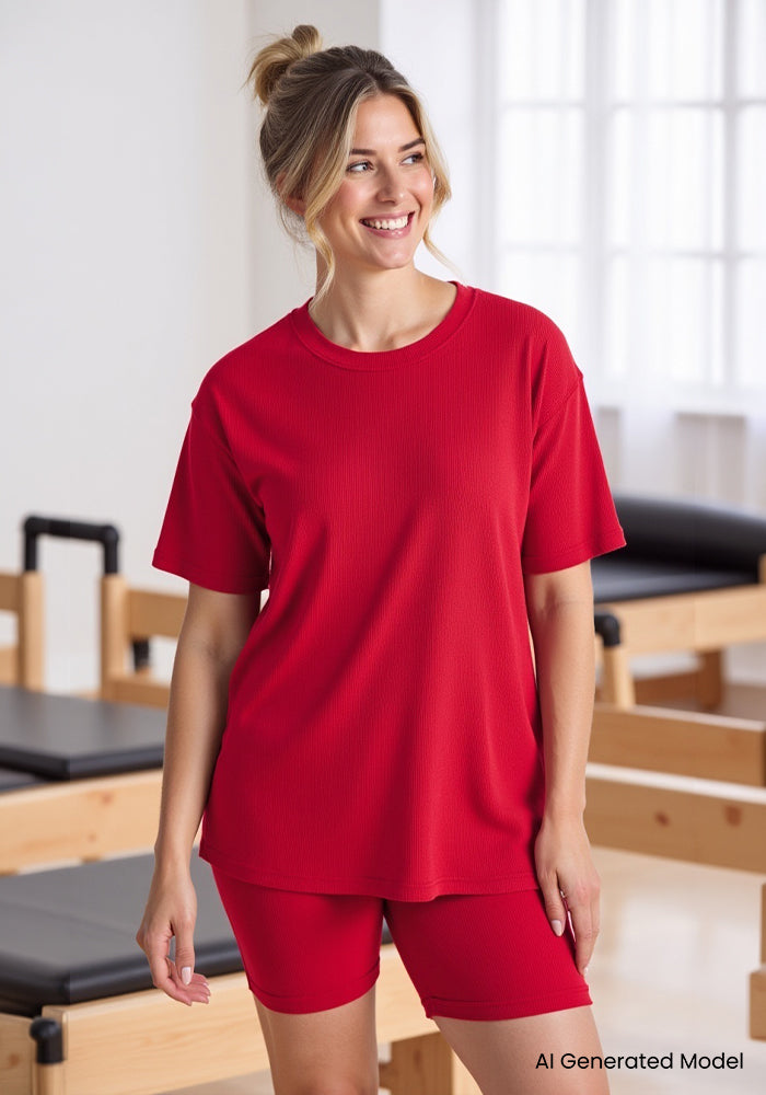Woman in the Woolx Vera Ribbed Tunic in Poppy Spark smiles in a bright room with Pilates equipment.