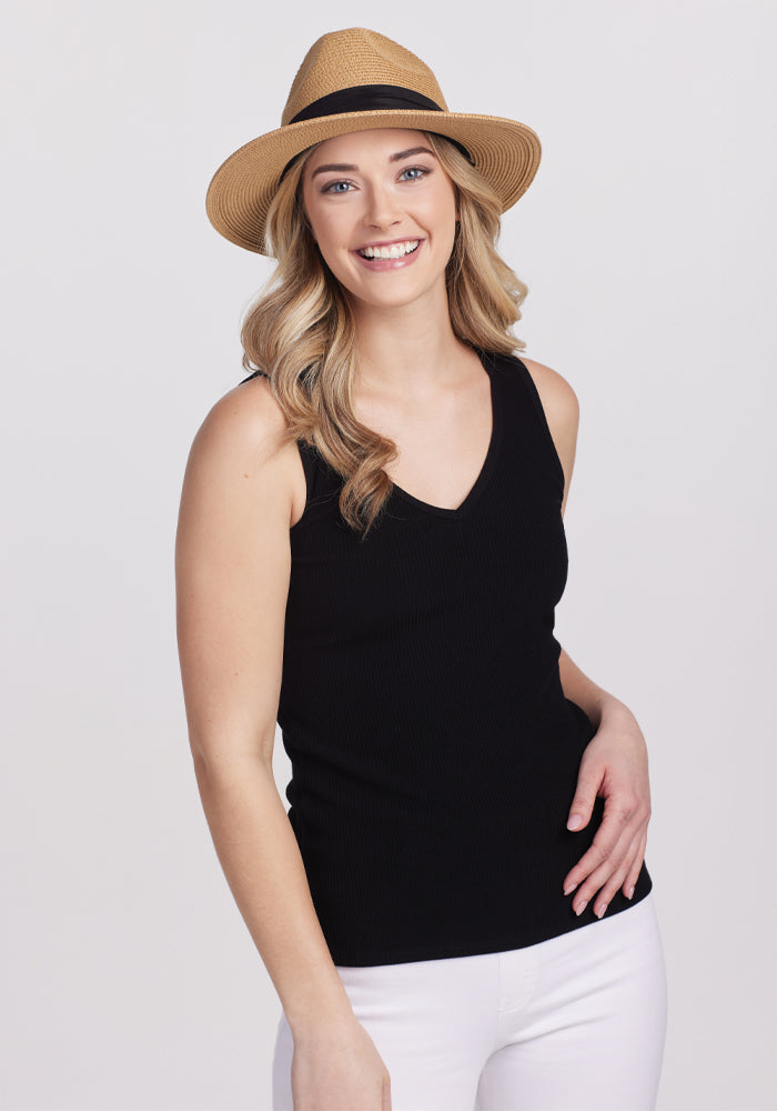 A woman in a Black Vivi Ribbed Tank by Woolx and a straw hat smiles at the camera, with long, wavy hair that captures capsule wardrobe vibes, against a plain light backdrop. | Ariana is 5’11”, wearing a size S