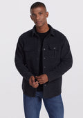 A man wearing the Woolx Wilder Shirt Jac in Carbon Black over a black t-shirt and blue jeans stands against a plain white background, exuding ultra-functional style as he looks directly at the camera. | Trell is 6’2”, wearing a size M