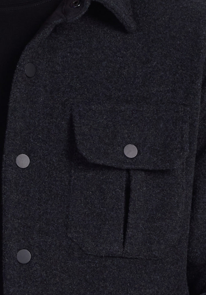 Close-up of the Woolx Wilder Shirt Jac in Carbon Black, crafted from Merino wool. This dark gray jacket features a flap pocket secured by a button and is adorned with black buttons along the front and collar. It embodies an ultra-functional design with a subtle textured appearance.