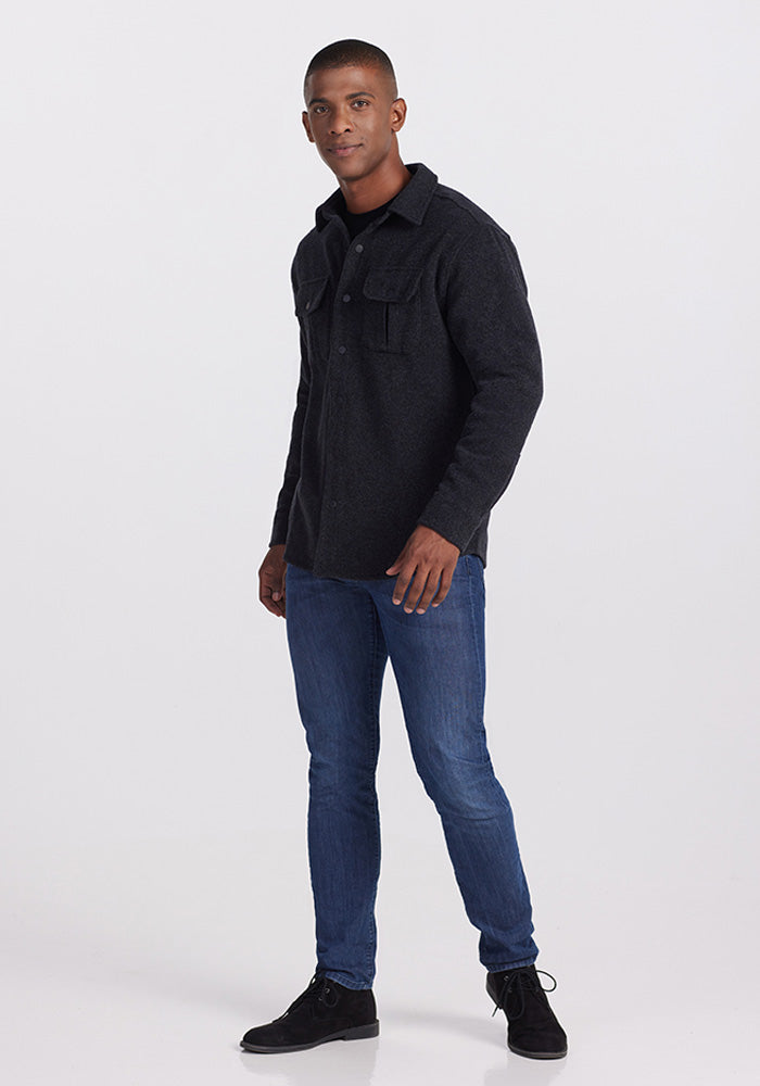 A person is standing against a plain background, showcasing the lumberjack style in a Wilder Shirt Jac in Carbon Black by Woolx, made from ultra-functional Merino wool. They pair it with blue jeans and black shoes, facing slightly to the left with a neutral expression.