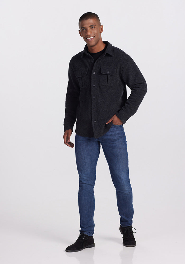 A person stands smiling in the Woolx Wilder Shirt Jac in Carbon Black, blue jeans, and black shoes against a plain white background. With one hand in their pocket and a slight angle to their stance, they exude ultra-functional comfort with their relaxed posture.