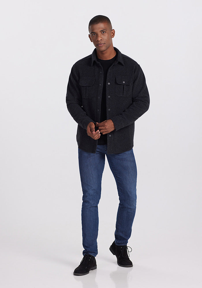 A person stands against a plain background, wearing the Wilder Shirt Jac in Carbon Black from Woolx over a black merino wool t-shirt, paired with blue jeans and black shoes. They are looking forward and holding their hands together in front.