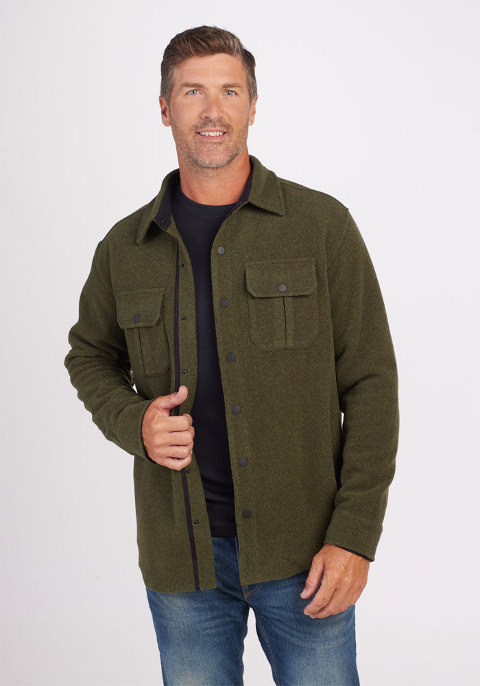 A man in a Woolx Wilder Shirt Jac in Dark Forest, a heavyweight button-up jacket crafted from Merino wool, is smiling at the camera. He pairs it with a black shirt and blue jeans. He has short hair and a beard, set against a plain white background.