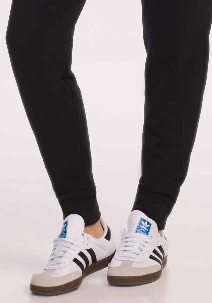 A person wearing Woolx Willa Joggers in black and white Adidas sneakers with black stripes stands on a light surface, with only the lower legs and feet visible.