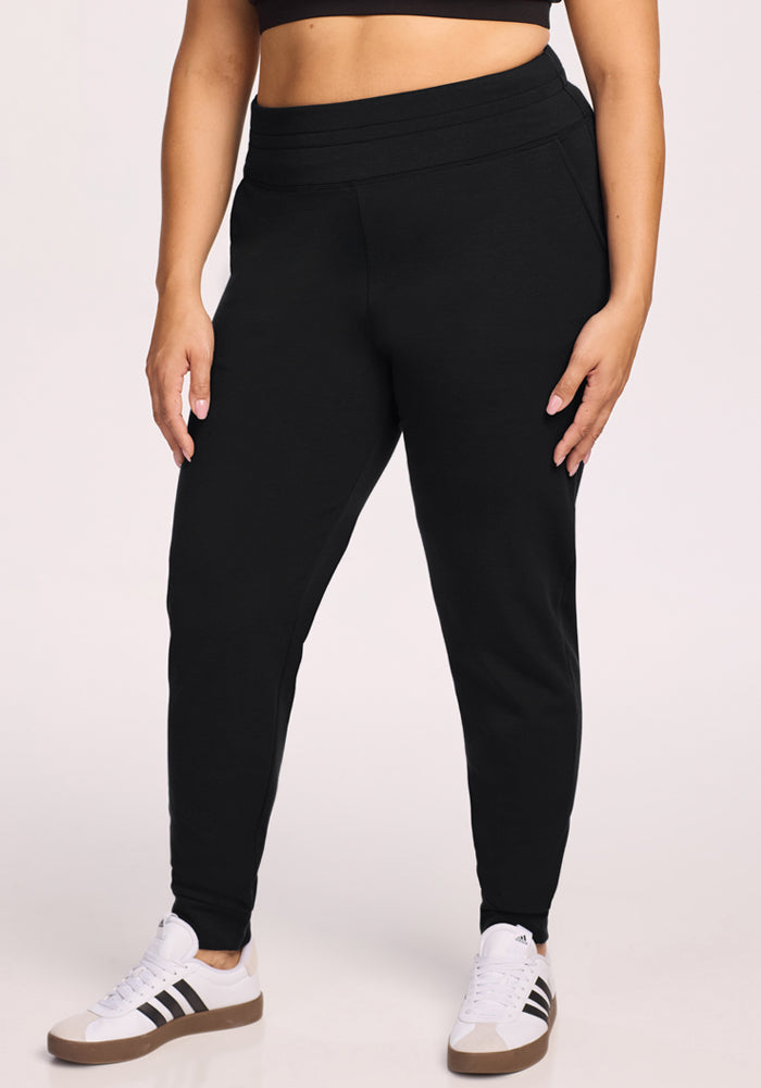 A person wearing Woolx Willa Joggers in black, paired with a black crop top and white sneakers with black stripes, stands against a light background with their hands relaxed by their sides. | Juliana is 5'10", wearing a size XL