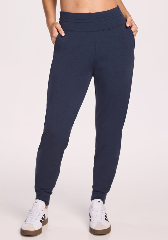 Someone stands against a plain background, hands in pockets, wearing Woolx Willa Joggers in Deep Navy and white sneakers with black stripes. | Ariana is 5'11", wearing a size S