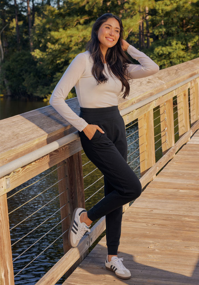 A woman with long dark hair stands on a wooden bridge by a lake, smiling. She wears a light long-sleeve shirt, Woolx Willa Joggers in Deep Navy, and white sneakers. Green trees and water are visible in the background.