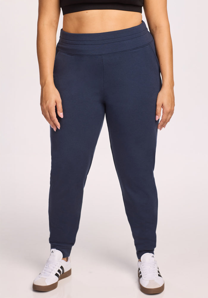 A person wearing Woolx Willa Joggers in Deep Navy, a black crop top, and white sneakers with black stripes stands against a plain light background. Only their body from shoulders to feet is visible. | Juliana is 5'10", wearing a size XL