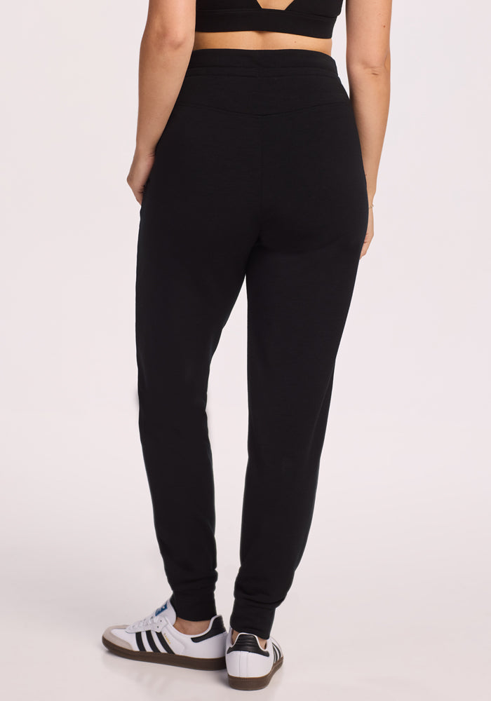 A person stands with their back to the camera against a plain light background, wearing Woolx Willa Joggers Petite in black, a black crop top, and white Adidas sneakers with black stripes.