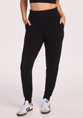 A person wearing Woolx Willa Joggers in black, paired with a black crop top and white sneakers with black stripes, stands with hands in pockets against a plain light background. | Saige is 5’7”, wearing a size XS