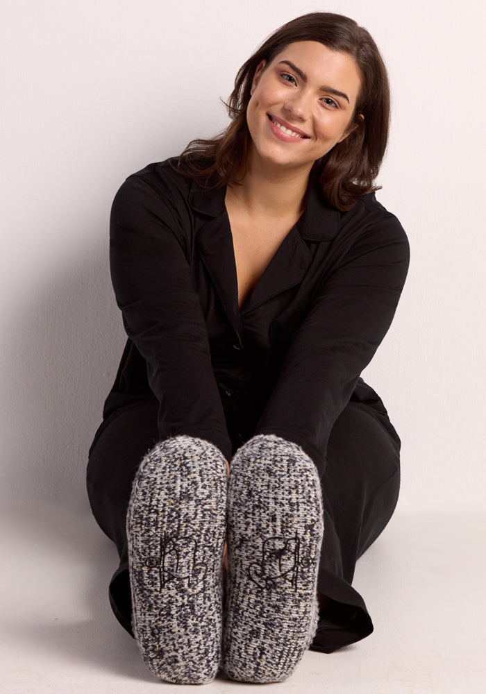 A smiling woman with brown hair in black attire sits on the floor, showing off the soles of her cozy Woolx Women's Gripper Slipper Booties in Speckled Stone toward the camera.