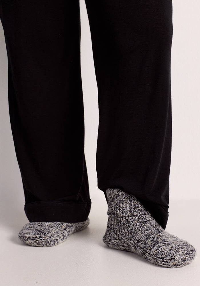 A person in black pants and grey marled knit socks stands on a light floor, ready to step into Woolx Women's Gripper Slipper Booties - Speckled Stone for extra warmth and comfort.