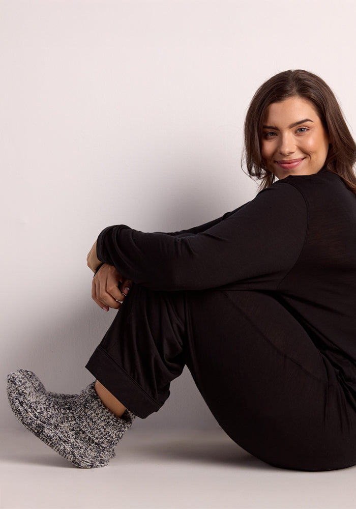 A woman with long brown hair in a black outfit wears Woolx Women's Gripper Slipper Booties in Speckled Stone, sitting on the floor with her knees up and smiling at the camera against a plain light-colored background.