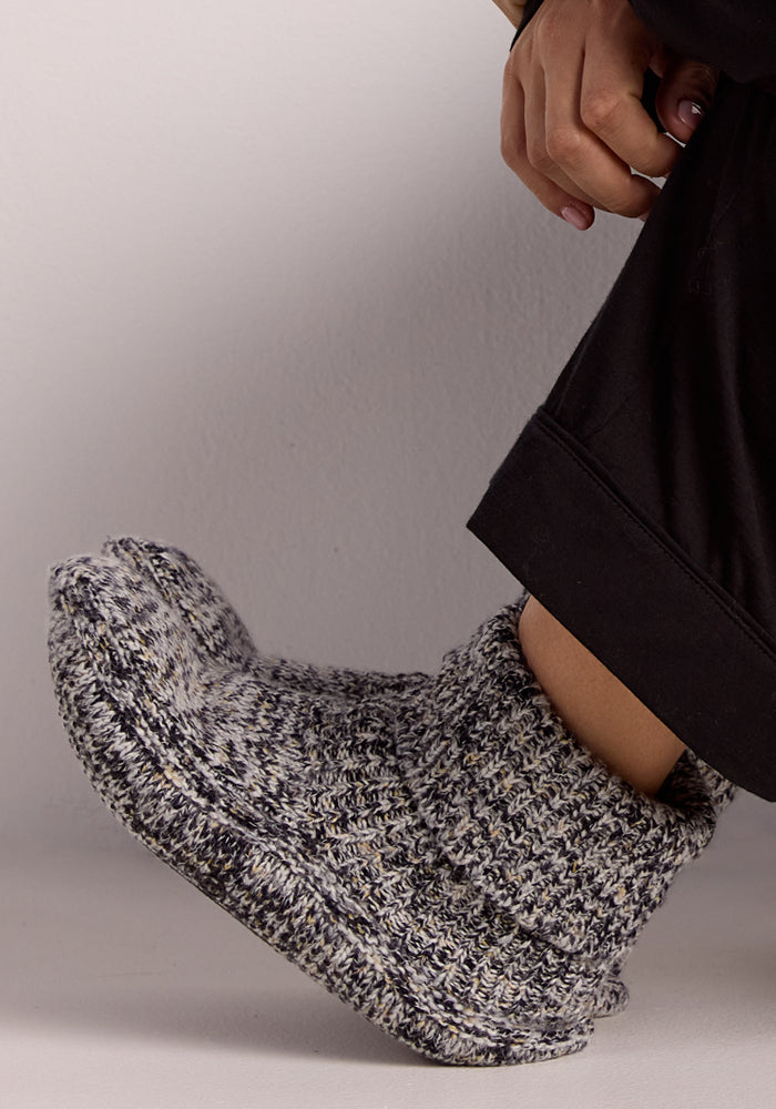 A woman in black pants lifts her foot while wearing Woolx Women's Gripper Slipper Booties in Speckled Stone, set against a plain light background.