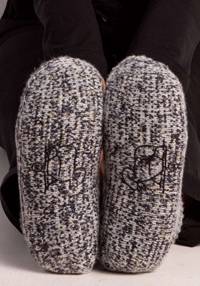 A person wearing Woolx Women's Gripper Slipper Booties in Speckled Stone, featuring cozy gray and black Merino wool with a decorative stitched sole pattern, sits with their feet together, soles facing forward.