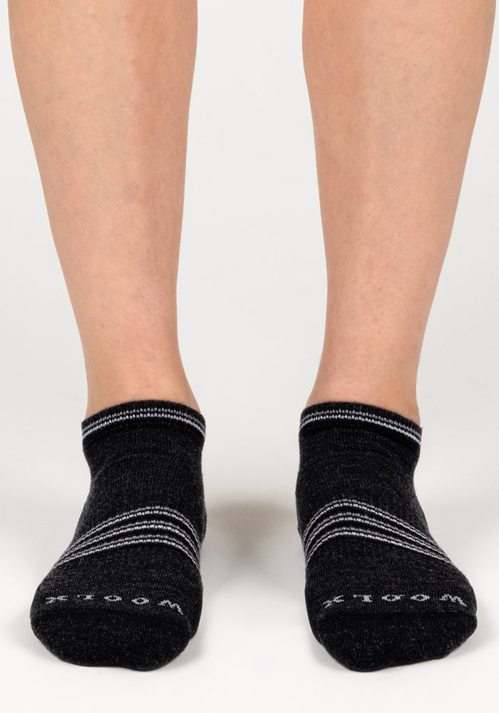 A close-up of a person wearing Woolx Womens Amelia No Show Socks in dark grey charcoal, featuring thin white stripes and "WOOL" on the toes, standing on a white surface.