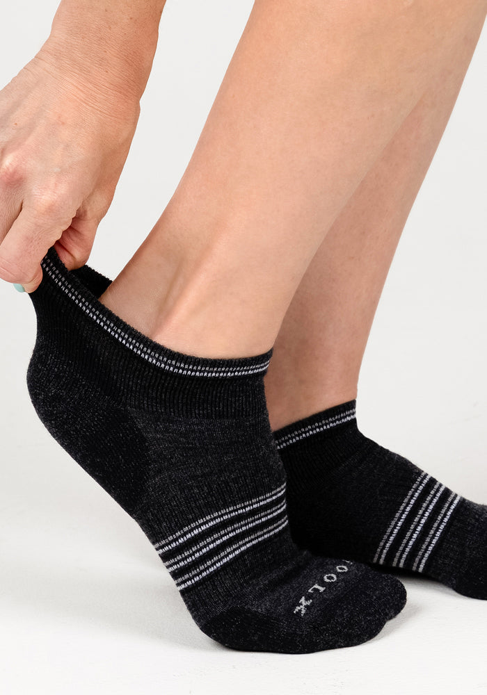 A person pulls on a Woolx Womens Amelia No Show Sock in dark grey charcoal. The matching sock is already worn on the other foot. Both socks feature white stripes, against a plain light background.