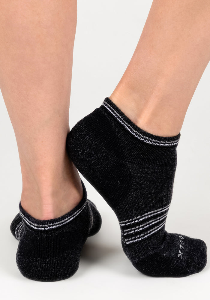 A close-up of two feet wearing Woolx Womens Amelia No Show Socks in Dark Grey Charcoal, with white stripes near the cuffs, standing on a white background—one foot is flat while the other rests on its toes.