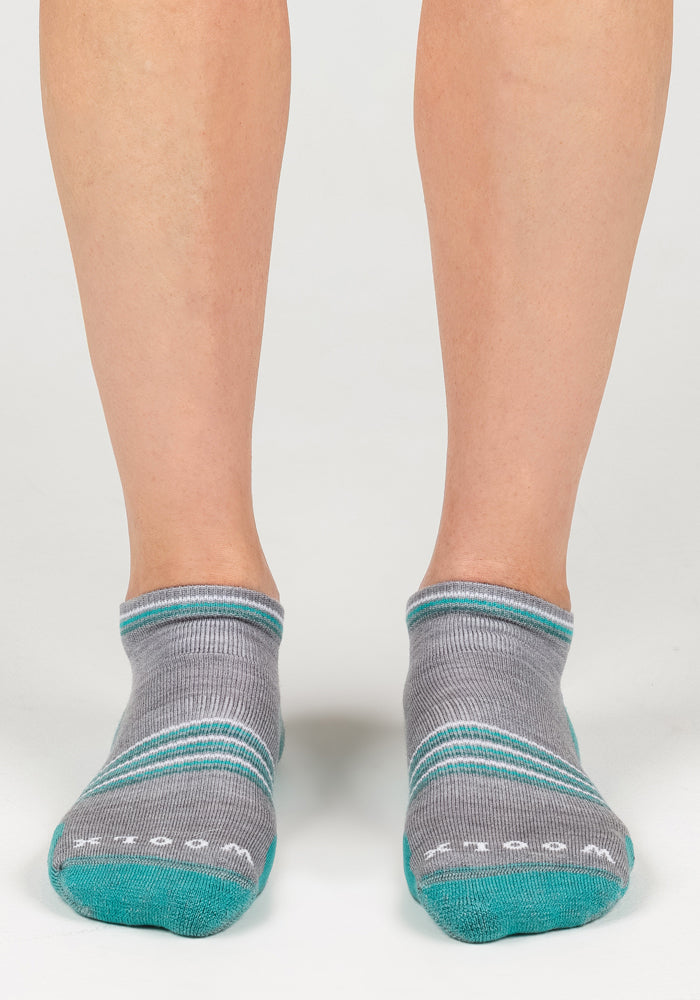 A pair of feet wear Woolx Womens Amelia No Show Socks in grey with teal and white stripes, teal toes, and white writing near the toes, standing on a white surface.