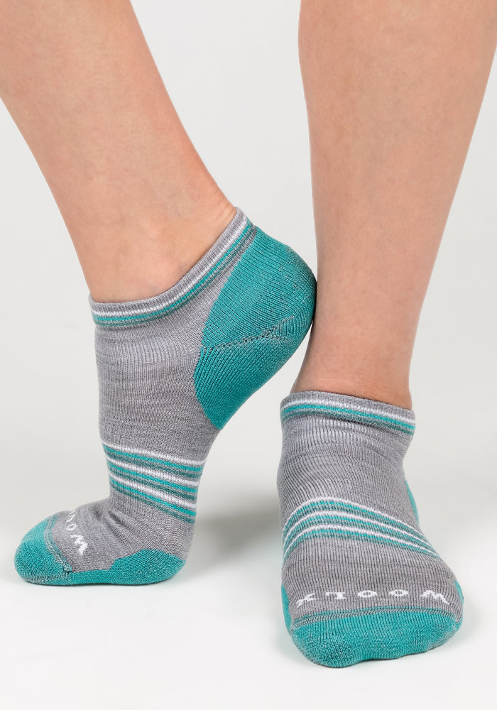 A close-up of two feet in Woolx Womens Amelia No Show Socks - Grey, featuring teal heels, toes, and stripes. One foot is slightly raised, showing the sole and side against a light background.