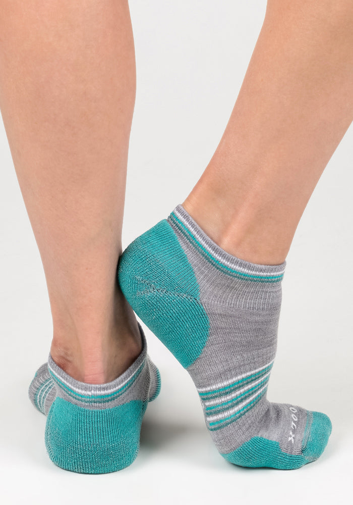 A pair of feet wearing Woolx Womens Amelia No Show Socks in grey, featuring teal heels and toes plus gray, teal, and white midfoot stripes, shown on a white background.