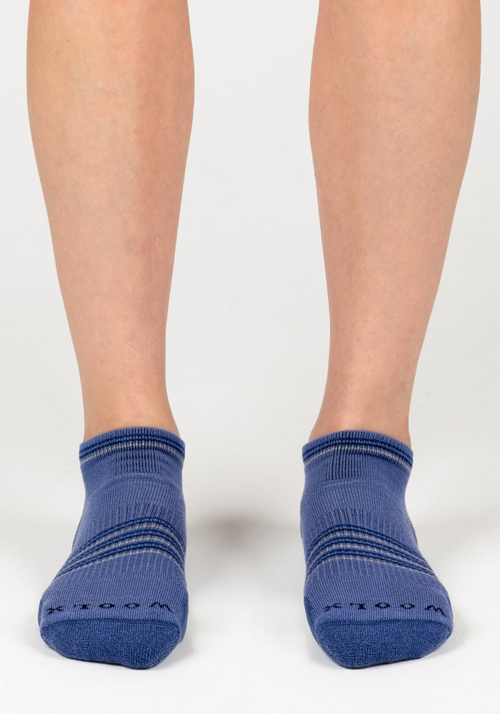 A close-up of a person’s lower legs and feet wearing Woolx Womens Amelia No Show Socks - Skyline, featuring blue, moisture-wicking fabric with subtle horizontal stripes, standing against a white background.