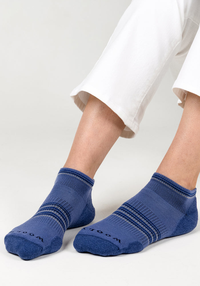 A person wearing white pants and Woolx Womens Amelia No Show Socks - Skyline sits with their feet crossed on a light-colored surface.