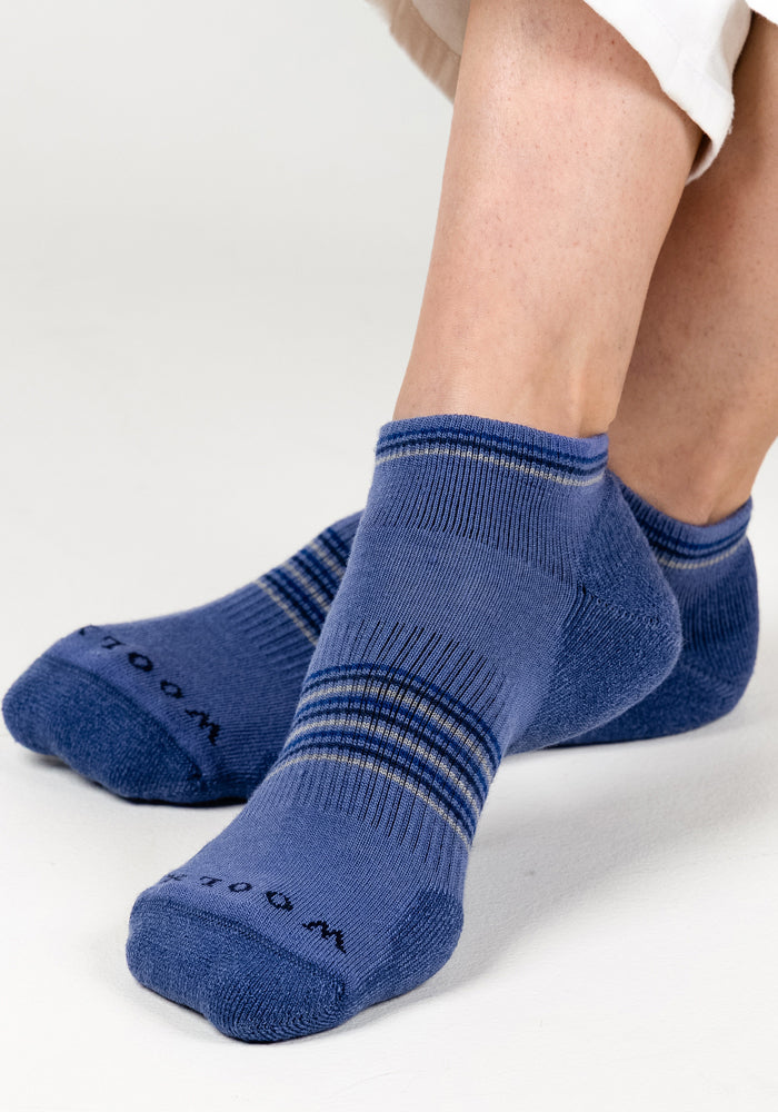 A close-up of a person wearing Woolx Womens Amelia No Show Socks - Skyline, featuring blue moisture-wicking fabric with thin horizontal stripes, paired with white pants and seated on a white surface.