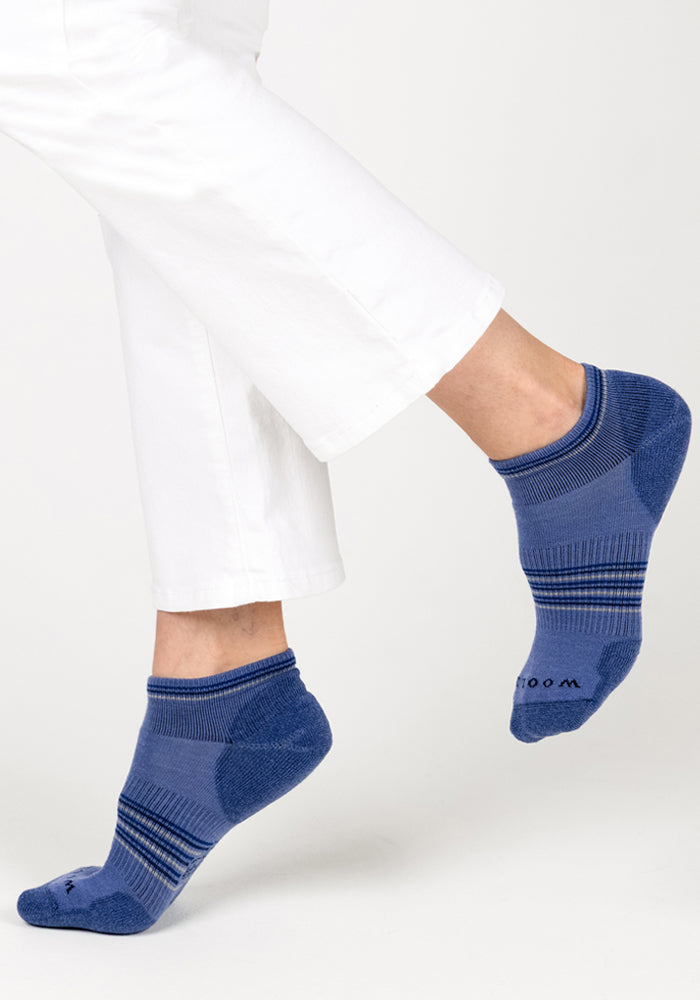 A person wears Woolx Womens Amelia No Show Socks - Skyline: blue ankle socks with dark blue heels, toes, and stripes, paired with white pants. Against a plain light background, these moisture-wicking socks provide comfort and style.
