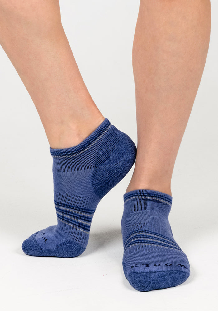 A woman stands on a light gray surface, one foot slightly lifted and crossed behind the other, wearing Woolx Womens Amelia No Show Socks - Skyline with blue moisture-wicking fabric and darker blue heels, toes, and stripes.