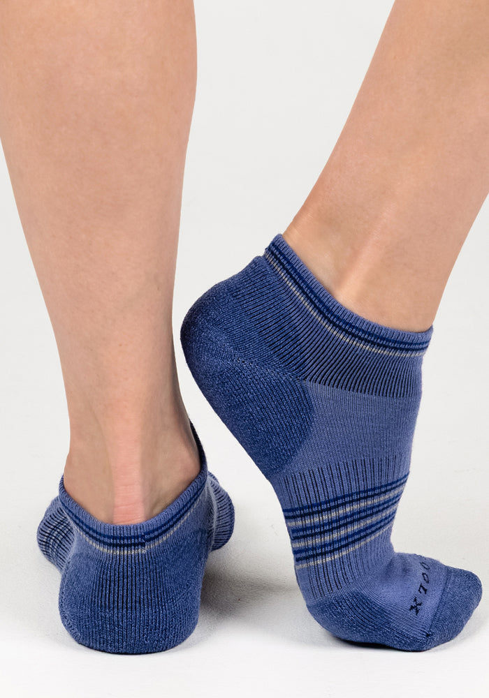 A close-up of a person wearing Woolx Womens Amelia No Show Socks - Skyline, featuring striped detailing and moisture-wicking Merino wool, standing on a white surface with one foot flat and the other raised to show the back and side.