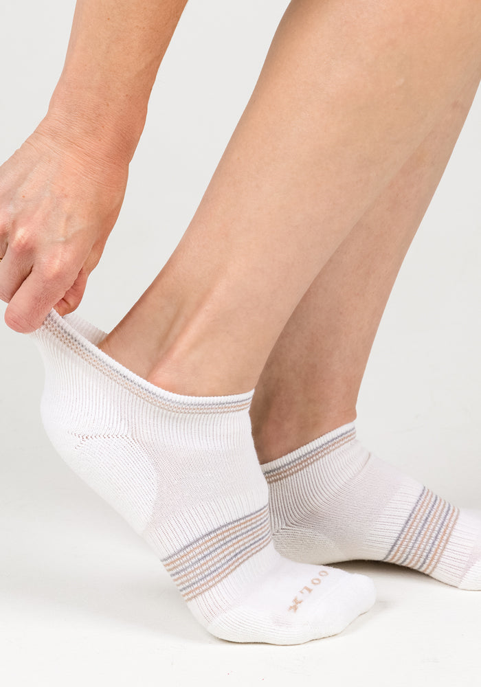 A person pulls on a Womens Amelia No Show Sock - White Lightning by Woolx, featuring moisture-wicking fabric and light gray/tan stripes, while already wearing the matching sock. The background is plain and light-colored.