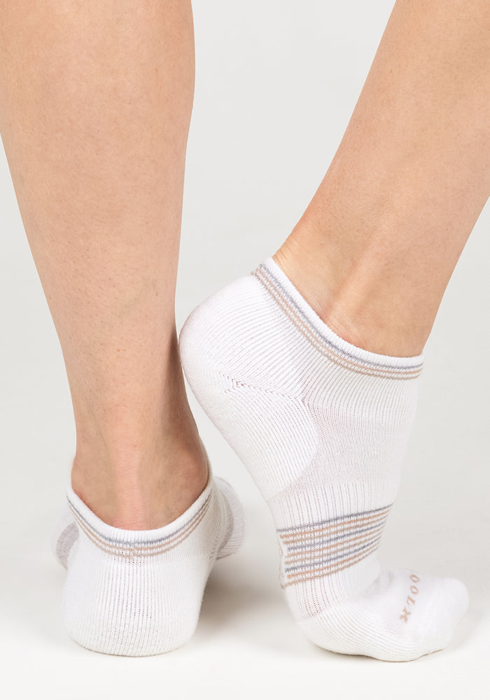 Close-up of two feet in Woolx Womens Amelia No Show Socks - White Lightning, featuring beige and gray heel stripes. One foot is flat while the other is on tiptoe, both on a white surface.