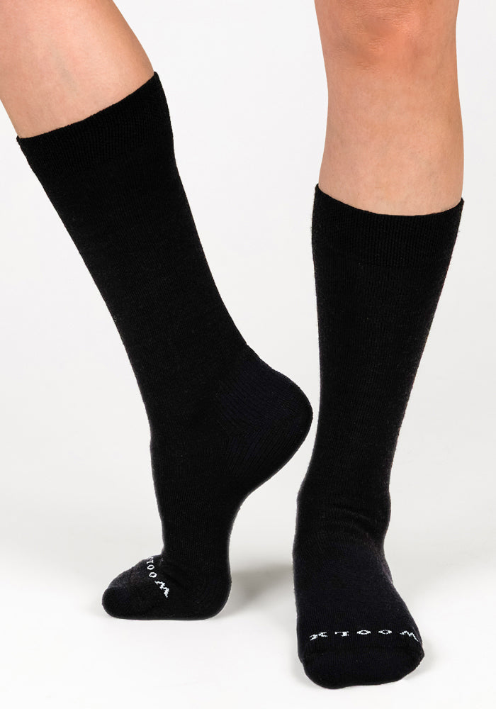 A person wearing Woolx Womens Chandler Mid Calf Socks in black stands on one foot with the other slightly raised. These lightweight, odor-resistant socks feature white toe text and are set against a plain, light background.