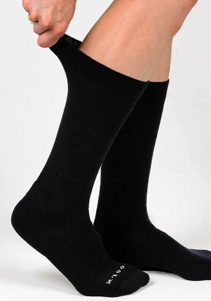 Someone is pulling up a pair of Woolx Womens Chandler Mid Calf Socks in black, showcasing their lightweight design against a white background.