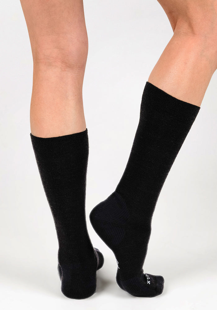 A person stands on a white surface, showing the back of their lower legs and feet wearing Woolx Womens Chandler Mid Calf Socks in Black. One foot is flat while the other heel is slightly raised.