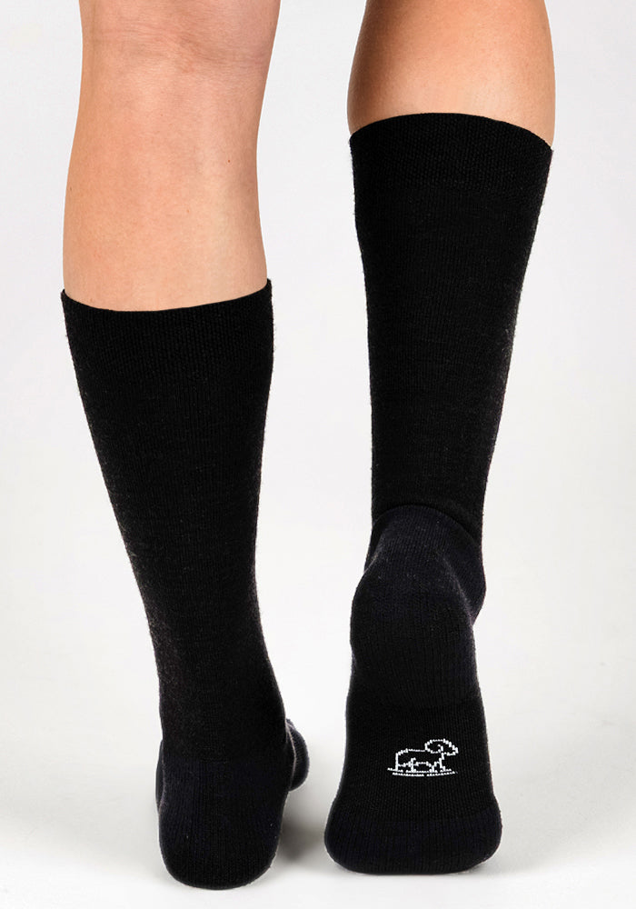 A person wears Woolx Womens Chandler Mid Calf Sock - Black, which are lightweight and feature a small white dog outline on the right heel. The photo shows the back of their legs against a plain, light background.