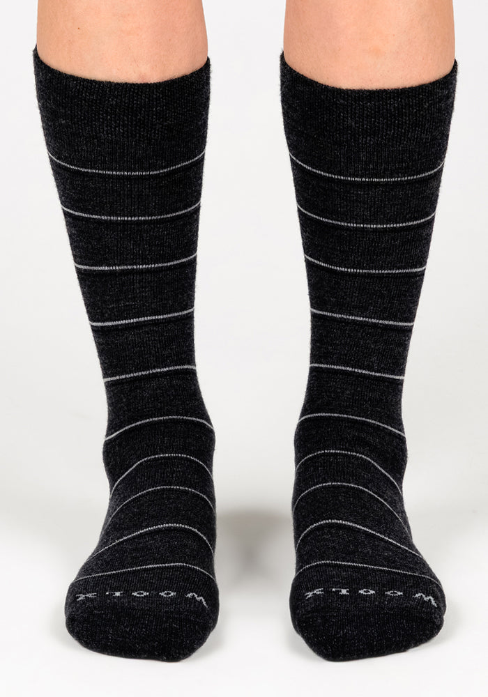 A pair of feet in Woolx Womens Chandler Mid Calf Socks in dark grey charcoal, featuring thin white stripes and "WOOL" on the toes; these lightweight Merino wool crew socks reach mid-calf.