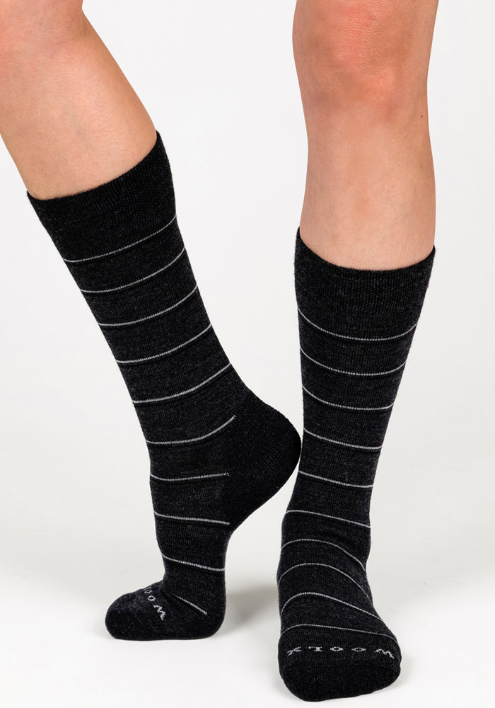 A person stands on a white background wearing Woolx Womens Chandler Mid Calf Socks in Dark Grey Charcoal, with one foot slightly ahead to display both side and front views of these odor-resistant Merino wool dress socks.