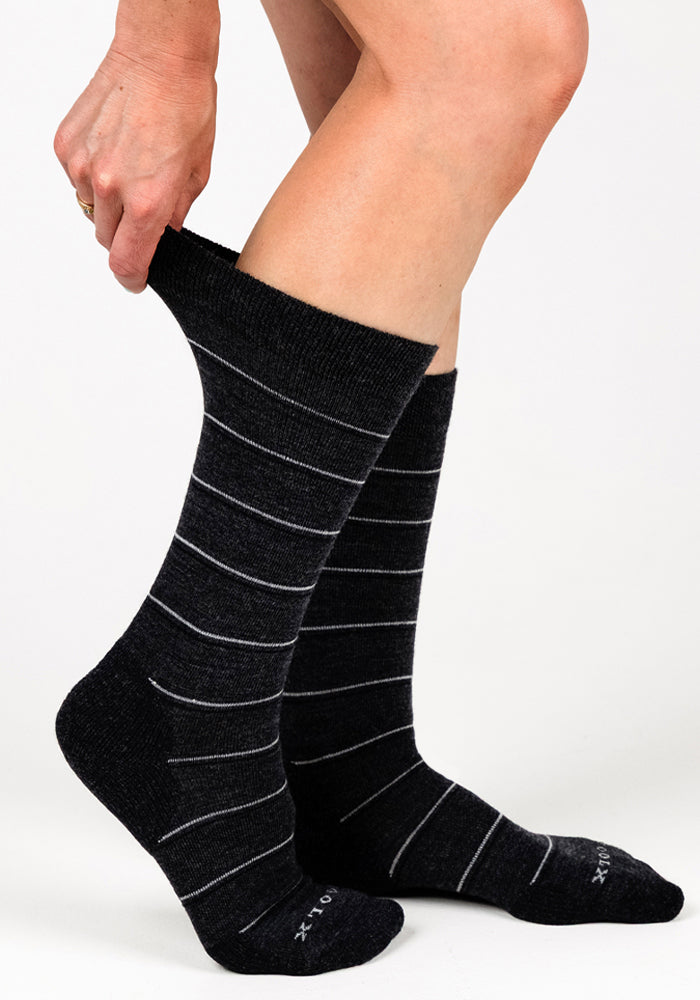 A person pulls up a Woolx Womens Chandler Mid Calf Sock in Dark Grey Charcoal, featuring thin white stripes, with a matching sock on the other foot against a plain white background.