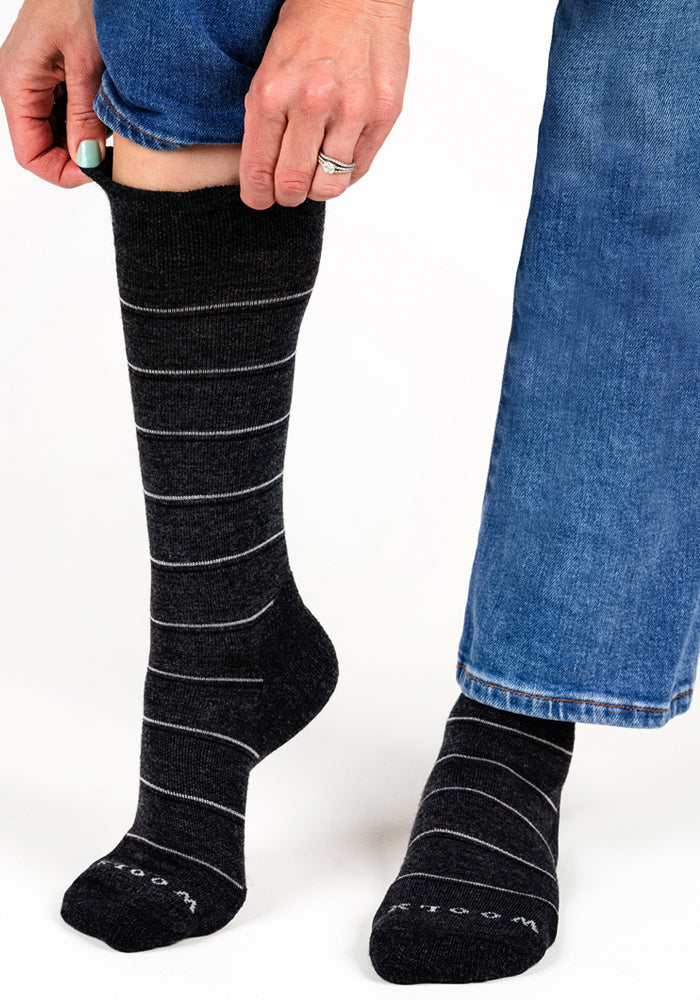 A person in blue jeans pulls up a Woolx Womens Chandler Mid Calf Sock in dark grey charcoal—an odor resistant Merino wool sock with thin white stripes and "Wool" near the toes. The other, matching sock rests on the ground.