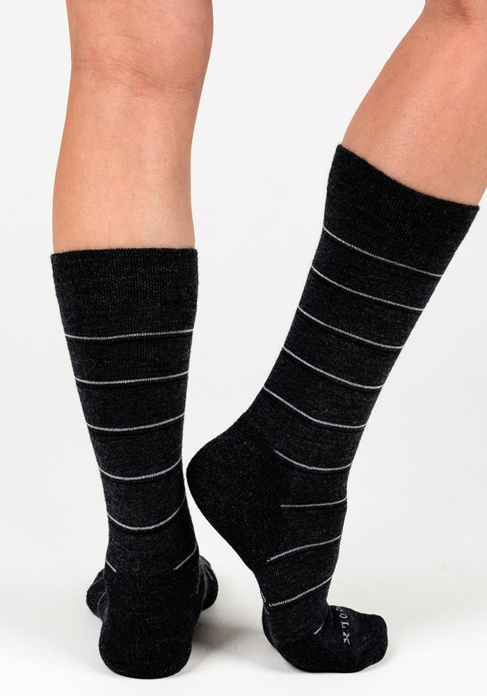 A person wears Woolx Womens Chandler Mid Calf Socks in Dark Grey Charcoal, featuring thin white stripes. One foot is flat, the other lifted onto its toes, showcasing the stylish and comfortable socks against a white background.